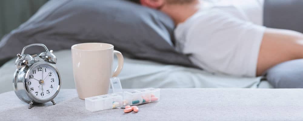 Sleeping Pills Lying Near Alarm-Clock While Man Sleeps In Bed